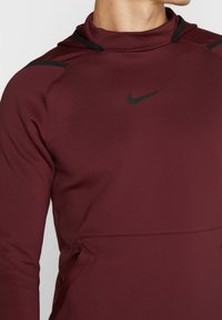 Maronfarvet Nike hættetrøje med en tætsiddende design, høj hals og sorte detaljer på skuldrene. Glat stof med en subtil glans. Med sidelommer.