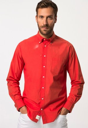 Camicia rossa a maniche lunghe con bottoni, realizzata in cotone, con taschino frontale e bottoni bianchi, indossata con pantaloni bianchi.