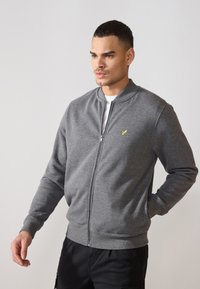 Pilkas zip-up megztinis iš minkštos medžiagos, su gumbuoto audinio rankovėmis ir apačia, su maža geltona emblema kairėje krūtinės pusėje.