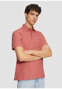 Rotes kurzärmliges Poloshirt mit strukturiertem Finish, einem Kragen und einer Brusttasche. Mit geradem Saum und seitlichen Schlitzern. Getragen mit hellblauen Jeans.