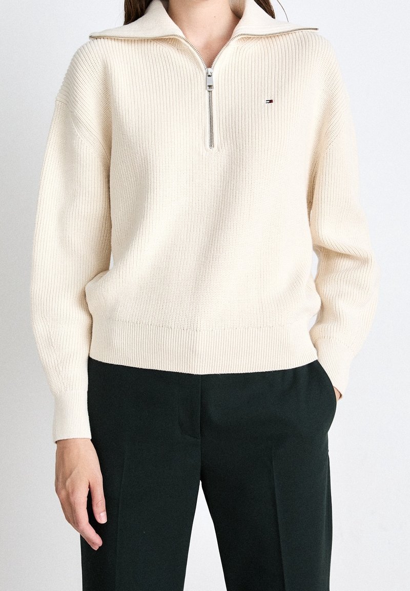Personne portant un pull en maille côtelée crème avec un col demi-fermeture éclair et un pantalon noir, debout devant un fond clair uni.