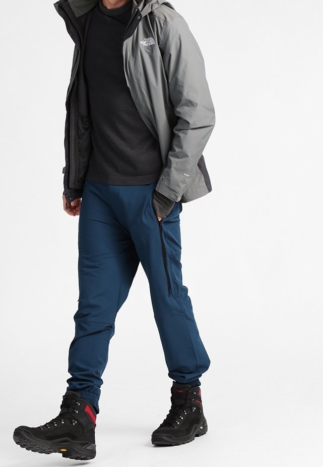 Homme portant une veste grise, une chemise noire, un pantalon bleu avec des poches zippées et des chaussures de randonnée noires marchant sur un fond blanc.