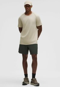 Camisa corta de manga corta texturizada en color beige, combinada con pantalones cortos verdes oscuros y zapatillas marrones. La gorra coincide con el color de la camisa. Diseño simple y atlético.