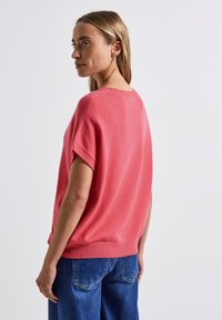 Pull en maille corail à manches courtes, avec un ourlet côtelé et une coupe décontractée. Texture douce, finition lisse, porté avec un jean en denim bleu.