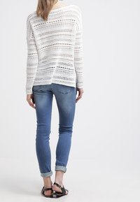 Pull en tricot blanc à mailles ouvertes avec une coupe décontractée, associé à un jean skinny bleu clair, retroussé aux poignets, et des sandales noires plates.