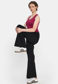 Bordowy top na ramiączkach, czarne szerokie spodnie i białe trampki z czarnymi paskami. Modelka stoi na jednej nodze, unosząc drugie kolano.