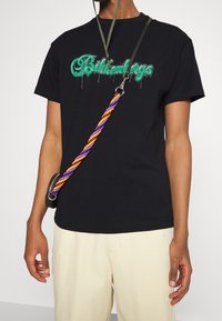 Czarna bawełniana koszulka z zielonym nadrukiem graficznym z efektem kapania, zestawiona z kremowymi spodniami o fakturze i torebką na kolorowym pasku.