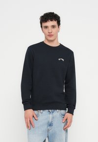 Tengerészkék pulóver hosszú ujjakkal, kerek nyakkivágással és bordázott mandzsettákkal. A mellkason apró fehér hímzett felirat található. Világos farmerral viselve.