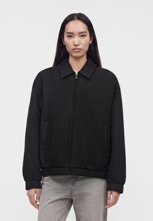 Frau mit langen dunklen Haaren, die eine schwarze Jacke mit Reißverschluss und hellgraue Jeans vor einem schlichten hellen Hintergrund trägt.