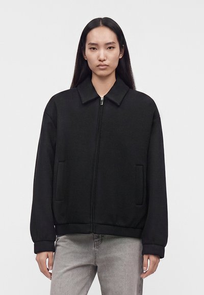 Femme aux longs cheveux foncés portant une veste zippée noire et un jean gris clair sur un fond clair uni.