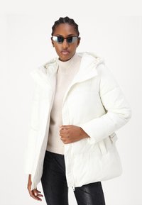 Hvid dunjakke med hætte, der har en frontlynlås og knaplukning. Båret over en lys beige sweater og sorte strukturerede leggings.