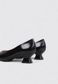 Zapatos de charol negros con punta redonda y tacones acampanados únicos. Superficie lisa con un acabado brillante y detalle de costura sutil.