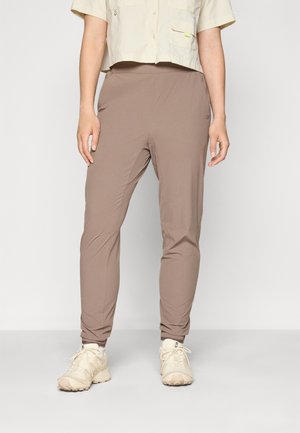 Frau trägt taupefarbene Slim-Fit-Jogginghose, cremefarbenes kurz geschnittenes Hemd mit Knöpfen und weiße Sportschuhe vor weißem Hintergrund.