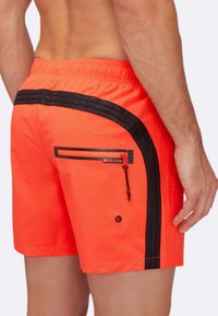 Uomo che indossa pantaloni da bagno arancioni vivaci con dettagli a strisce nere e una tasca con zip sul retro, mostrato dalla vita a metà coscia.