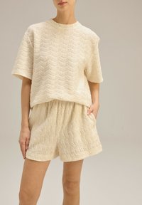 Kremowy, teksturowany strój składający się z bluzki z krótkim rękawem i dopasowanych szortów w falisty wzór. Materiał ma miękkie, dotykowe wykończenie.