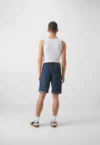 Denim šortky v tmavomodrej farbe s prevrátenými lemami, skombinované s bielym topom bez rukávov. Minimalistický dizajn, obsahujúci dve zadné vrecká a kožený štítok s logom značky.