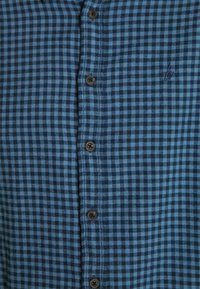 Camisa de cuadros azul y negra con botones, hecha de una tela de textura suave; presenta botones marrones y un pequeño logo bordado.