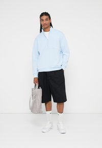 Sweatshirt de zíper azul claro, calções pretos, ténis brancos e bolsa cinza. Apresenta mangas longas, punhos canelados e um corte descontraído.