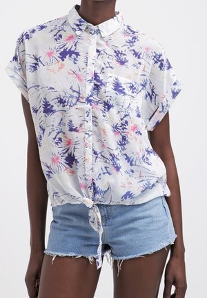 Woman wearing a short-sleeve white button-up shirt with purple and pink floral print tied at the waist and light blue denim shorts.