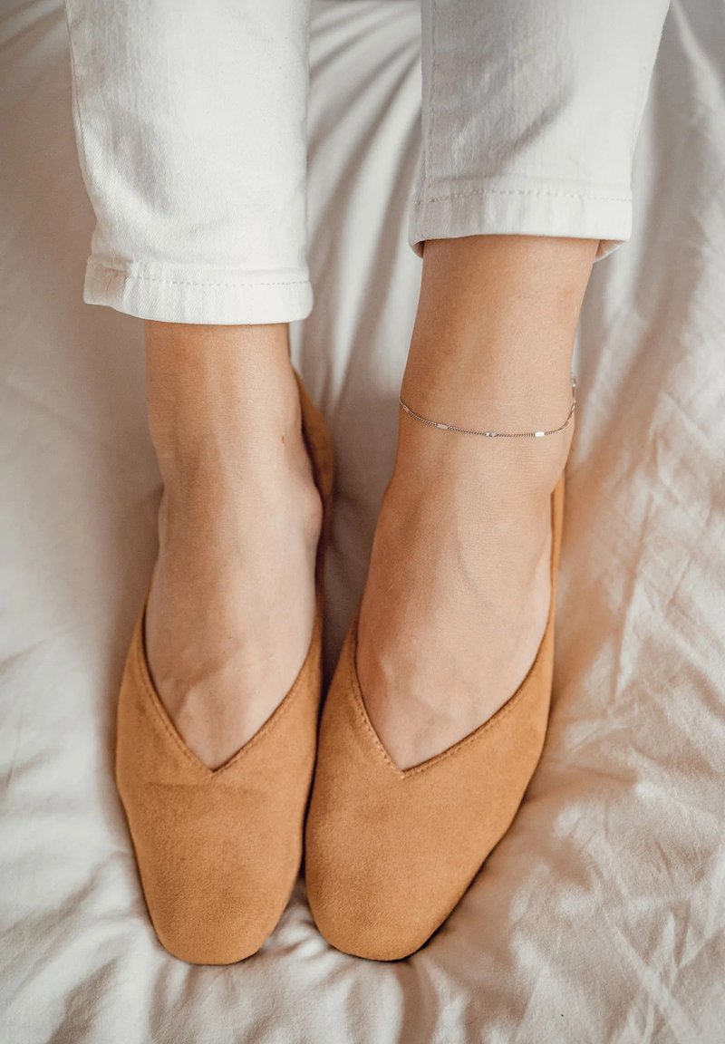 Ballerine in suede tan con design a V appuntito, morbide al tatto e dal profilo basso, abbinate a pantaloni bianchi a 3/4, su una superficie di colore chiaro.