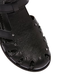 Sandalo in pelle nera con borchie metalliche, dettagli ritagliati e cinturino regolabile alla caviglia.