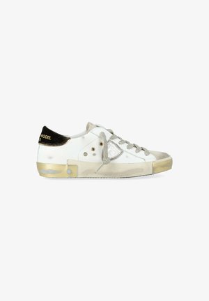 Sneaker bassa bianca con accenti beige, lacci grigi, patch metallico del logo sul lato e tab nera sul tallone con testo dorato.