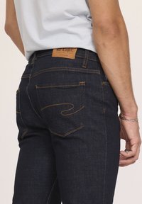 Jeans en denim bleu foncé avec une coupe slim, dotés de coutures orange, de deux poches arrière et d'une étiquette en cuir marron à la taille.
