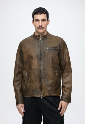 Homme aux cheveux courts et foncés avec une moustache, portant une veste en cuir marron avec des poches zippées et un pantalon noir sur un fond uni.