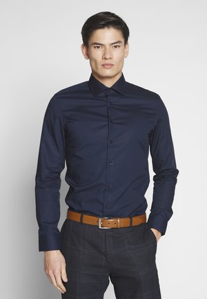 Homme portant une chemise bleu marine boutonnée avec une ceinture marron et un pantalon à carreaux foncé, debout avec une main dans la poche devant un fond uni.