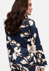 Granatowa bluzka z długimi rękawami, z białymi i złotymi wzorami kwiatowymi. Gładki, błyszczący materiał podkreśla wzór i fakturę.