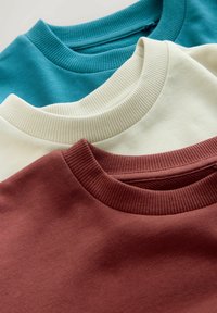 Tričko s okrúhlym výstrihom v teal, krémovej a burgundskej farbe. Hladká textúra s ribovanými výstrihmi. Každé tričko je naskladané a čiastočne sa prekrýva.