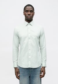 Šviesiai žalios spalvos ilgomis rankovėmis marškiniai su standartiniu apykakliu, siauru kirpimu ir lygiu audiniu, derinami su šviesiai mėlynomis džinsinėmis kelnėmis.