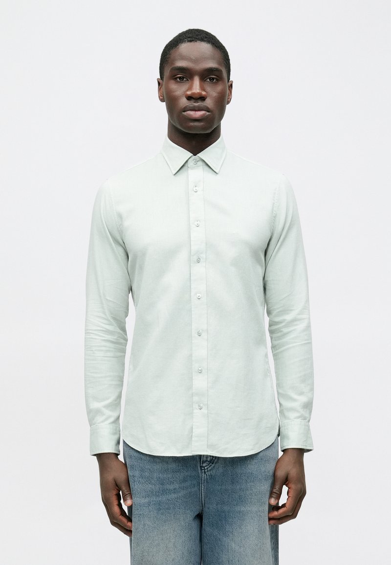 Šviesiai žalios spalvos ilgomis rankovėmis marškiniai su standartiniu apykakliu, siauru kirpimu ir lygiu audiniu, derinami su šviesiai mėlynomis džinsinėmis kelnėmis.