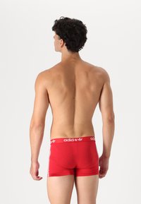 Rote Boxer-Briefs aus elastischem Stoff, mit einem weißen Streifendesign und einem Markenbund. Sicht von hinten, Modell nicht vollständig sichtbar.