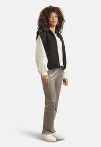 Schwarze ärmellose Weste über einer weißen bestickten Bluse mit langen Ärmeln, kombiniert mit glänzenden silbernen Hosen und weißen Sneakers. Einfaches und modernes Design.