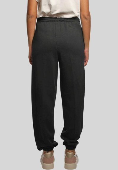 Persona che indossa pantaloni jogger neri con vita elastica e sneaker beige, in piedi davanti a uno sfondo grigio uniforme.