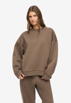 Jeune femme portant un sweat-shirt marron ample et un pantalon assorti, debout avec les mains près de l'ourlet du sweat-shirt, fond neutre.