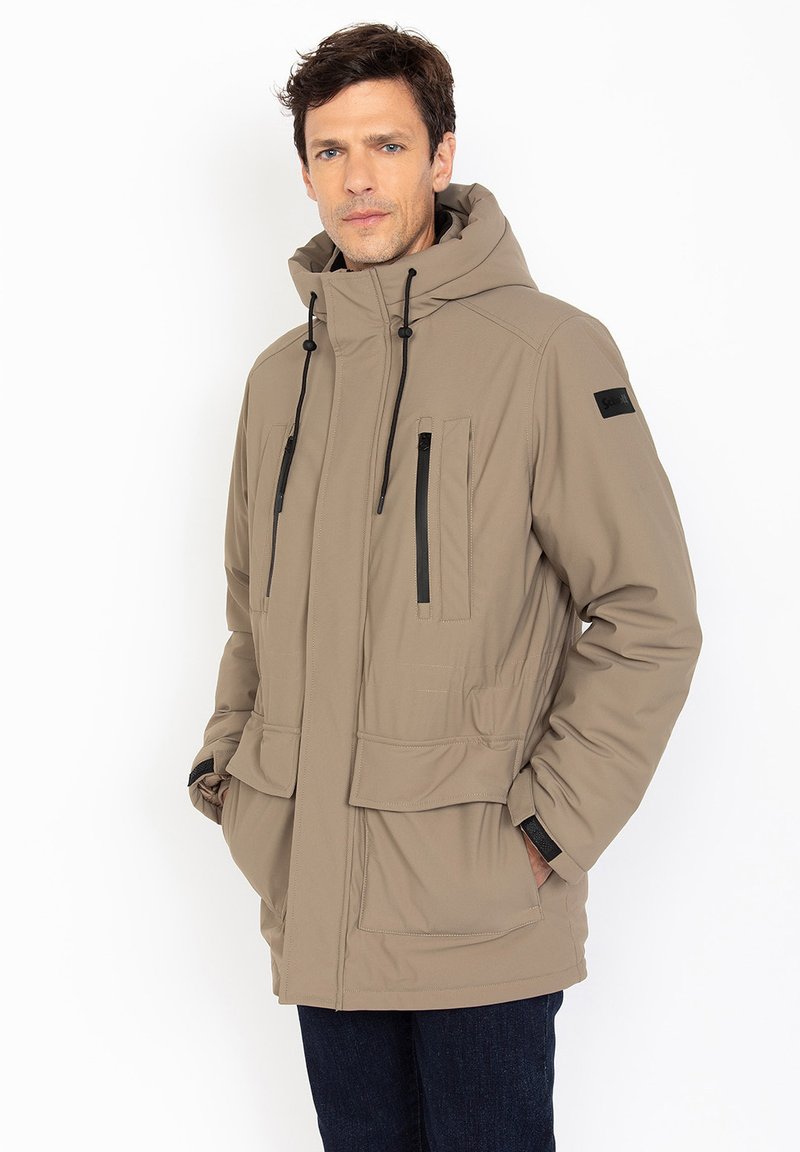 Chaqueta acolchada beige con capucha, bolsillos frontales y acentos en negro. Cuenta con cierre de cremallera y cordones ajustables. Textura suave.