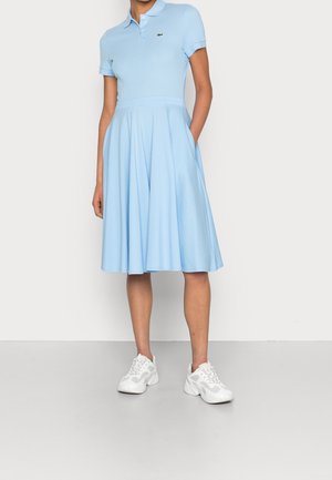 Femme portant une robe polo à manches courtes bleu clair avec une jupe évasée et des baskets blanches, debout avec une main dans la poche, devant un fond uni.