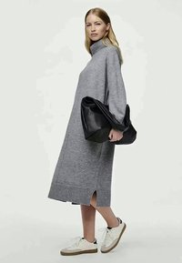 Γυναίκα με γκρι πουλόβερ φόρεμα και λευκά αθλητικά παπούτσια, κρατώντας μια μεγάλη μαύρη δερμάτινη τσάντα, στέκεται μπροστά από ένα απλό φόντο.