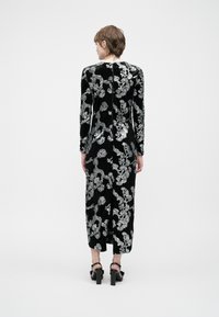 Pessoa vestindo um vestido de veludo preto com bordado floral prateado, de costas para a câmera, e usando sandálias de salto alto pretas em fundo simples.