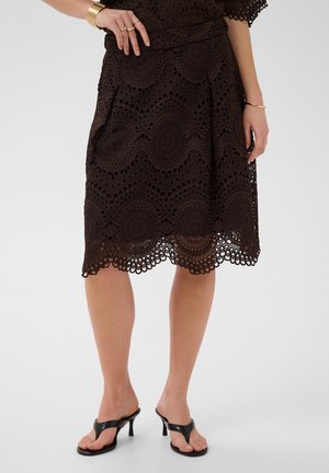 Jupe midi en dentelle noire avec ourlet festonné, portée avec des sandales à talons noires ainsi que des bracelets et bagues dorés aux mains.