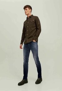 Rjava srajca z gumbi in dvema žepoma na prsih, v kombinaciji s temno modrimi slim-fit kavbojkami in črnimi gležnjarji. Gladka tekstilna površina.