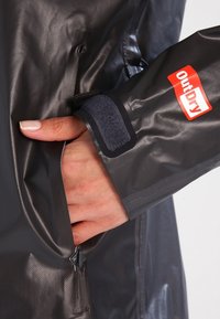 Vandtæt grå jakke med en tekstureret overflade, der har et synligt OutDry-logo, lynlåslommer og en velcro-rem ved manchetten.