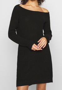 Černé pletené šaty s odhalenými rameny, dlouhými rukávy, ribbovanou texturou a mírně oversize střihem, končící nad koleny.