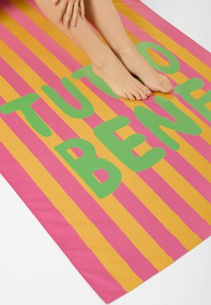 Barfüße und Hände ruhen auf einem pink-orange gestreiften Handtuch mit großen grünen Buchstaben, die „TUTTO BENE“ schreiben.