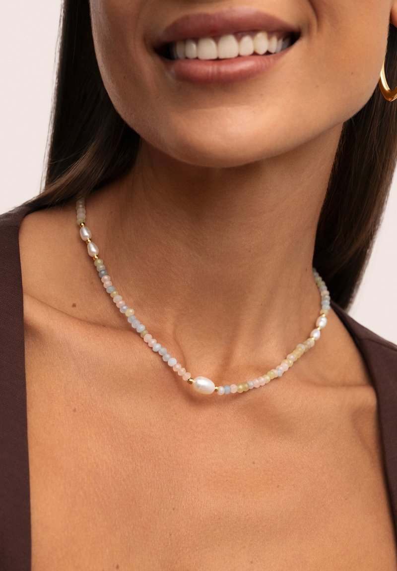 Een glimlachende vrouw met lang haar draagt een kralenketting met kleurrijke stenen en een enkele parel, tegen een neutrale achtergrond.