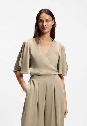 Femme aux longs cheveux foncés porte un chemisier beige style cache-cœur avec des manches larges et un pantalon plissé taille haute assorti, debout sur fond blanc.