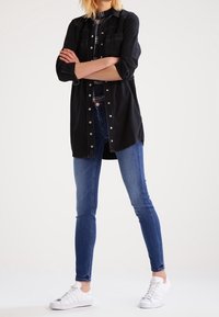 Camisa de mezclilla negra con detalles de botones, mangas largas y dos bolsillos en el pecho, combinada con jeans ajustados azules y zapatillas blancas.