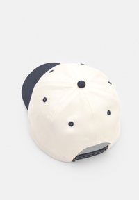 Casquette en coton blanche avec visière bleu marine. Design à six panneaux avec des accents de couture et fermeture arrière ajustable à pression.